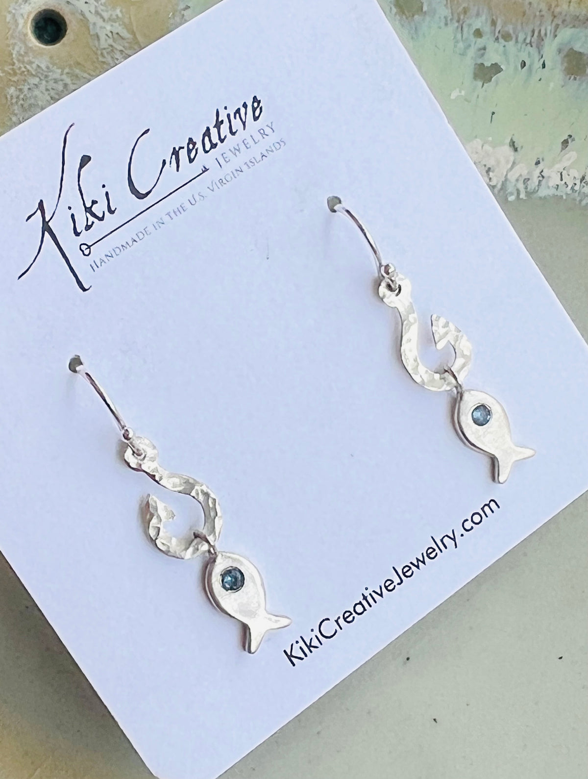 A pair of silver fish drop earrings with small dark green stones, displayed on a white Kiki Creative jewelry card.
