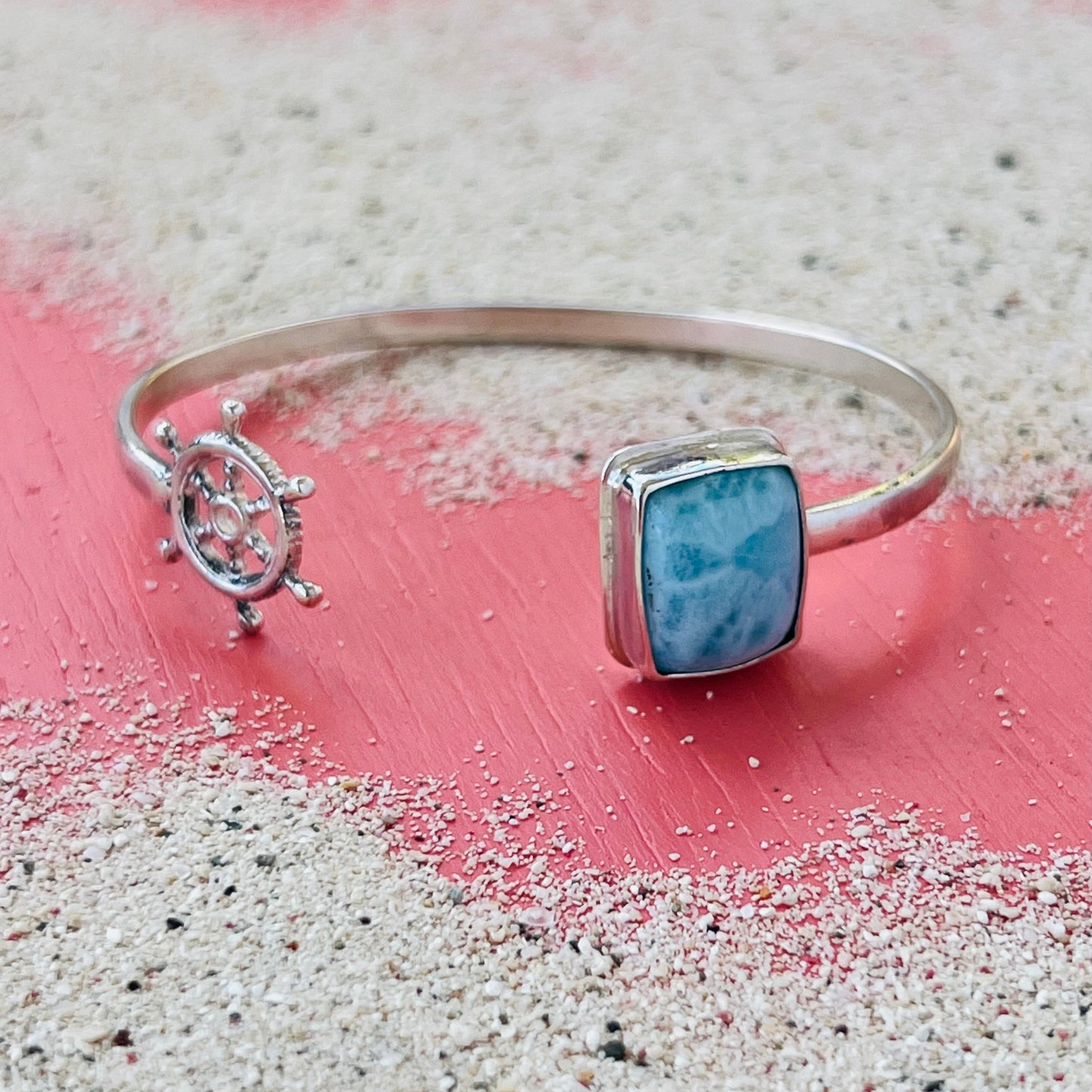 Silver bracelet with a blue stone and ship wheel charm on a sandy surface