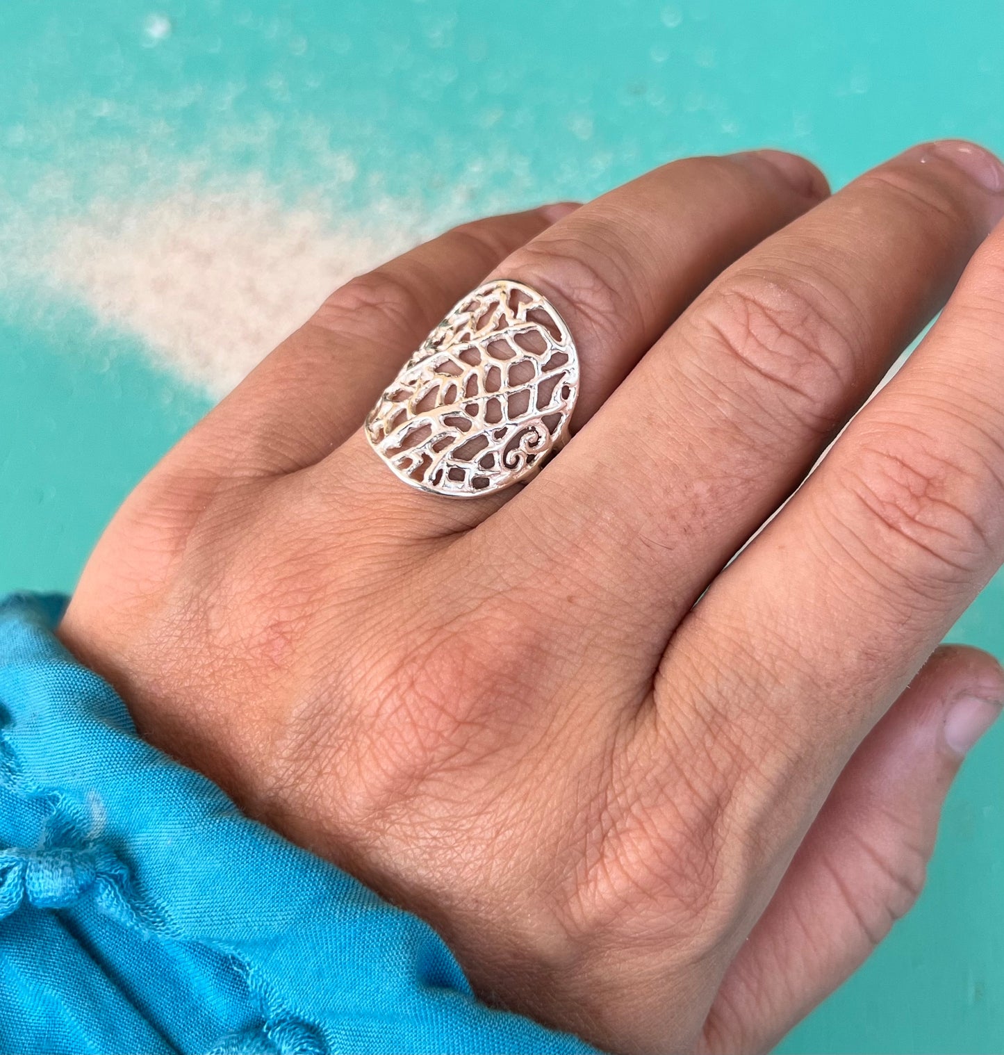 Hand wearing a silver lace ring against a turquoise background