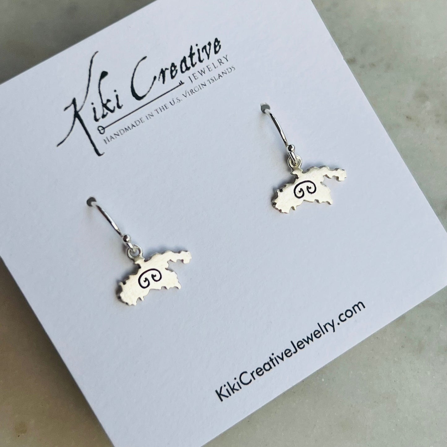 A pair of silver drop earrings shaped like the state of Michigan with a spiral symbol inside, displayed on a white Kiki Creative jewelry card.