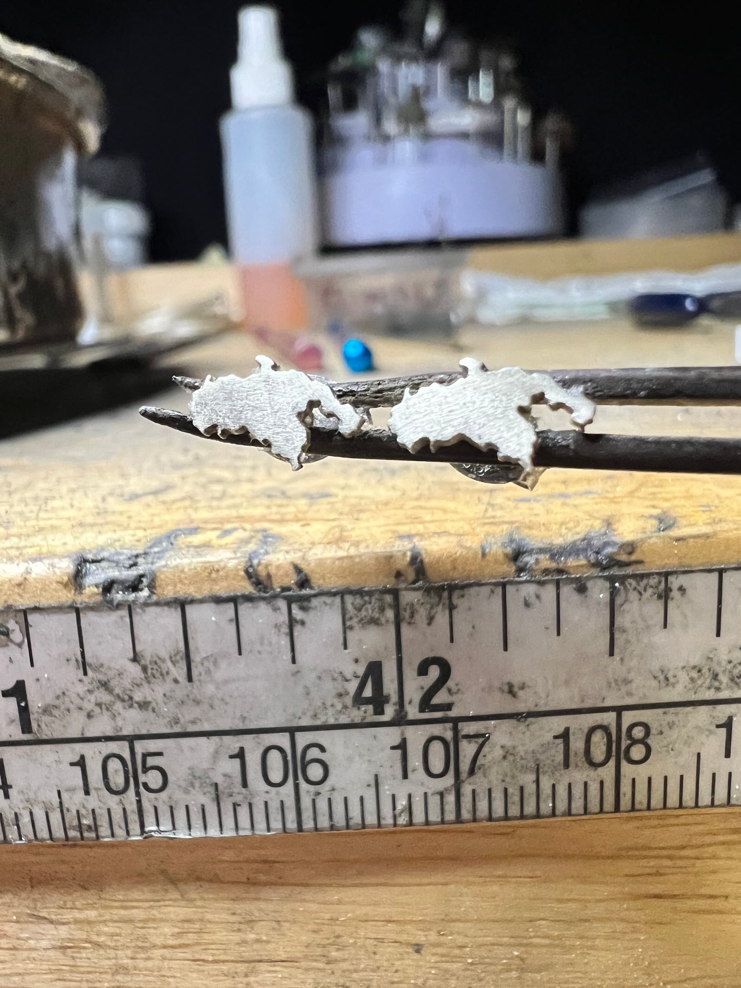 Two small silver-colored earring pieces held on forceps above a metal ruler on a wooden workbench.