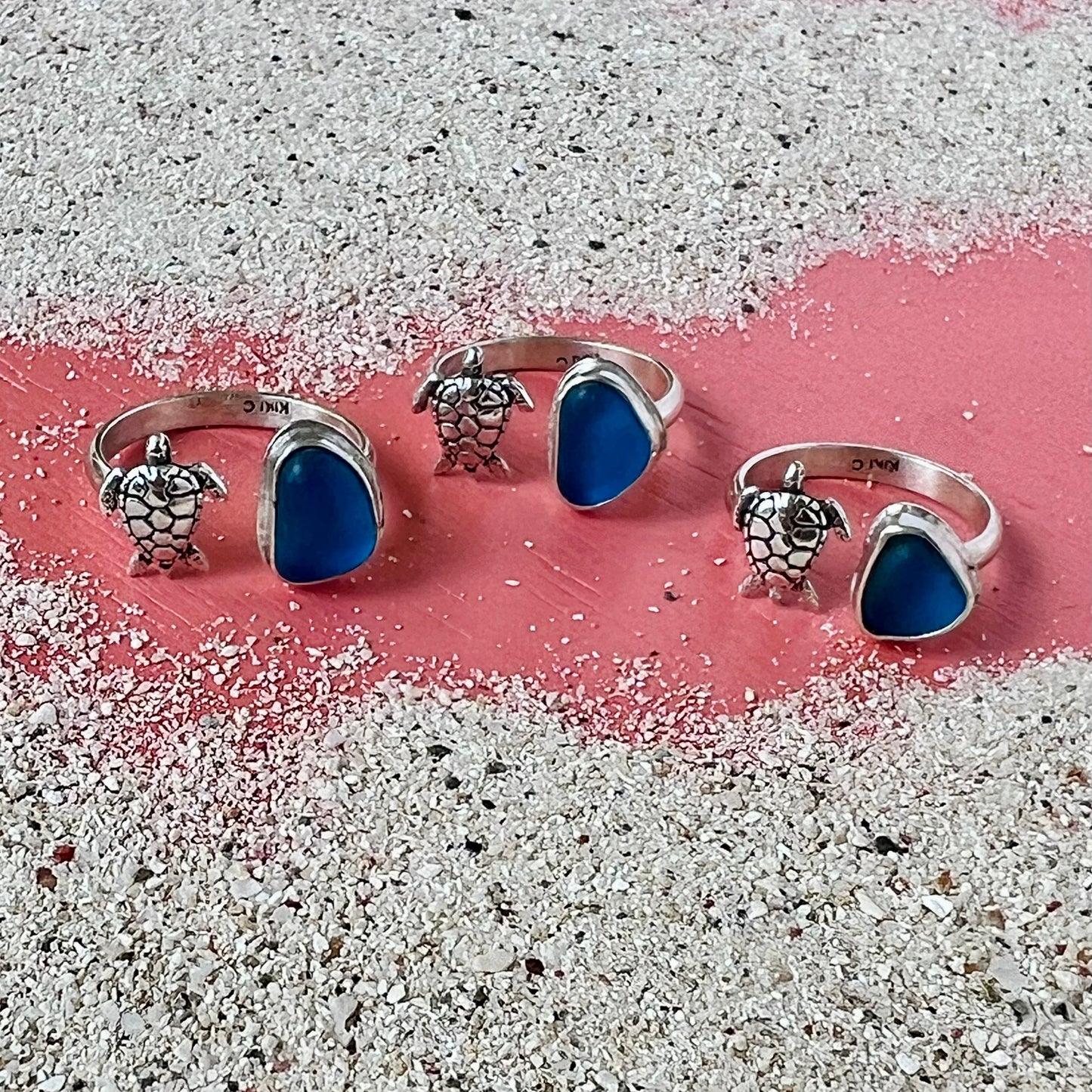 Three silver rings with blue sea glass and turtle designs on a sandy surface.