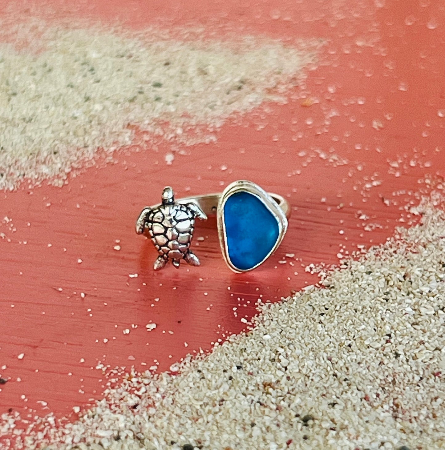 Silver turtle charm and blue sea glass ring on a sandy surface