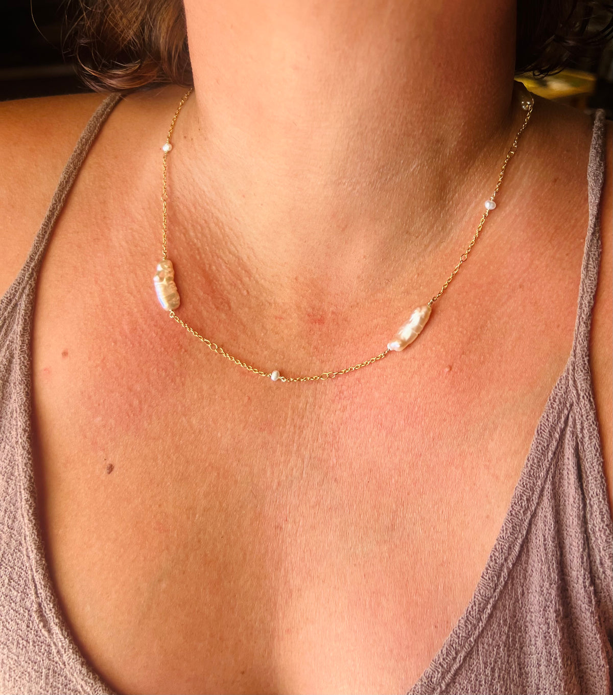 A close-up of a person's neck and chest wearing a delicate gold chain necklace with three irregular white baroque pearls and small clear stones.