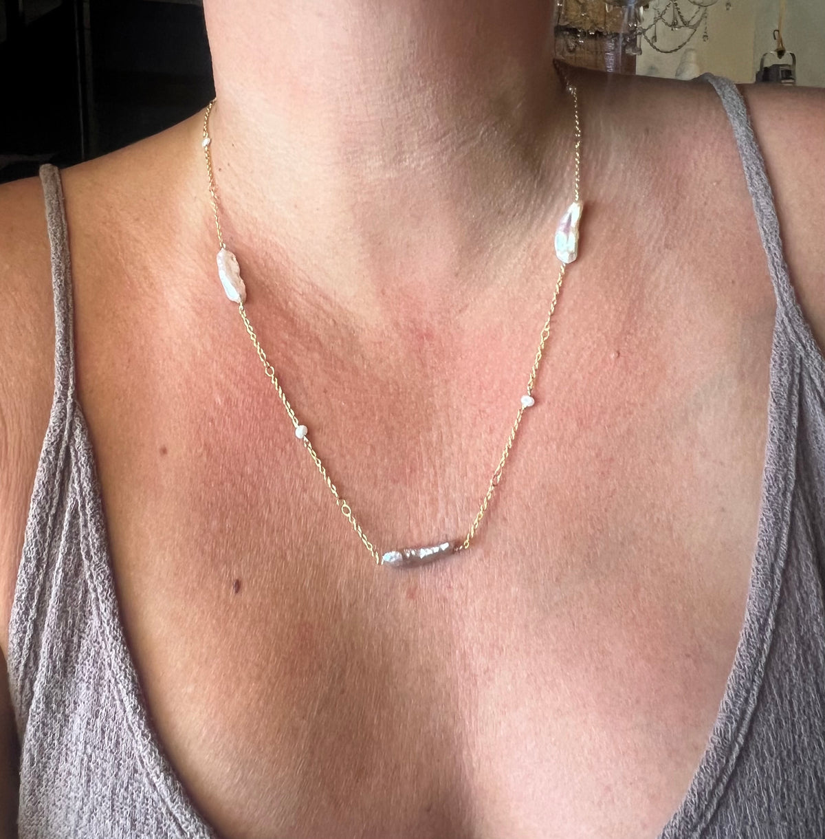 A close-up of a person wearing a delicate gold chain necklace with irregular white baroque pearls and small silver beads, styled with a gray tank top.