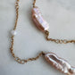 A gold chain necklace featuring two large, irregular light pink baroque pearls and a small white pearl bead, laid out on a light gray marble surface.