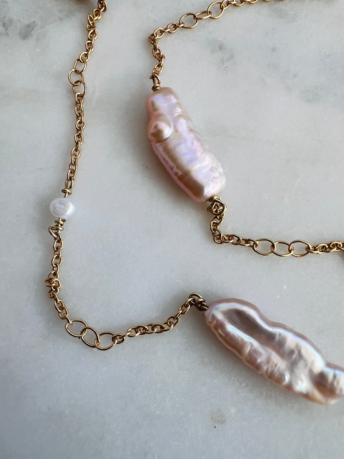 A gold chain necklace featuring two large, irregular light pink baroque pearls and a small white pearl bead, laid out on a light gray marble surface.