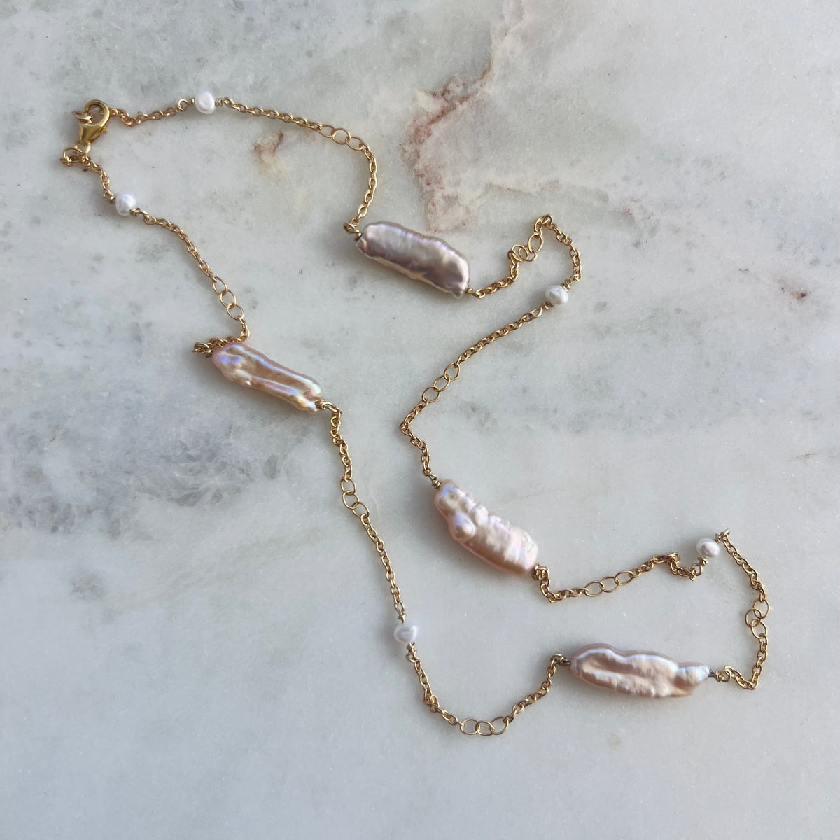 A delicate gold chain necklace featuring irregular baroque pearls in soft pink and cream tones, accented with small white pearls, laid out on a light gray marble surface.