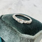 A silver band ring with emerald, oval and round-cut sapphire stones, displayed on a dark velvet jewelry cushion against a light marble background.