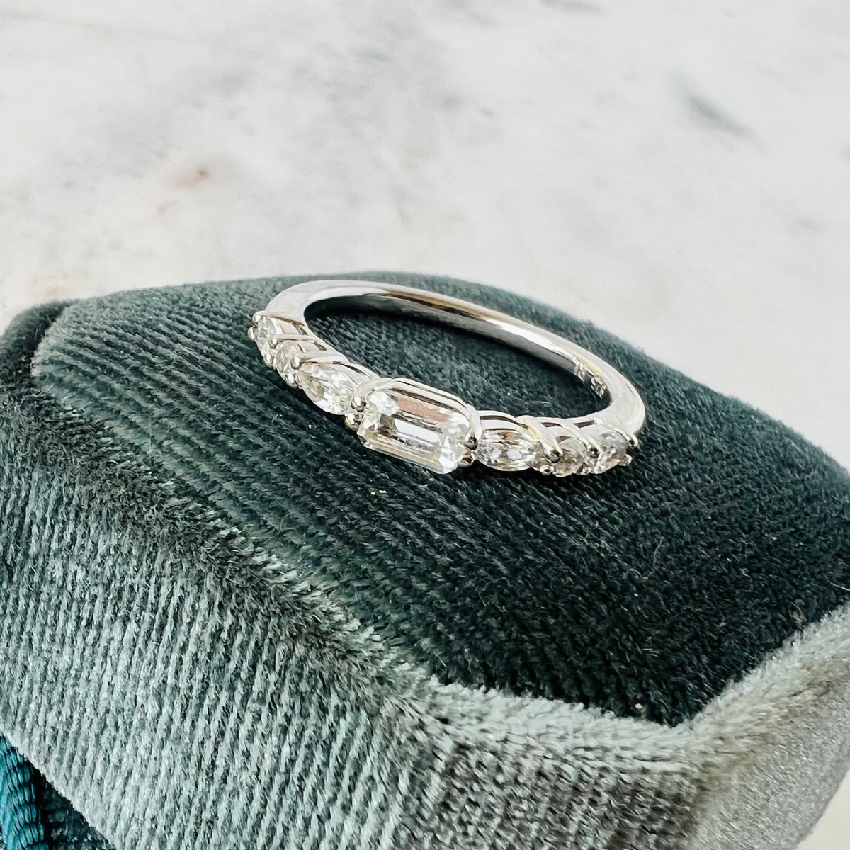 A silver band ring with emerald, oval and round-cut sapphire stones, displayed on a dark velvet jewelry cushion against a light marble background.