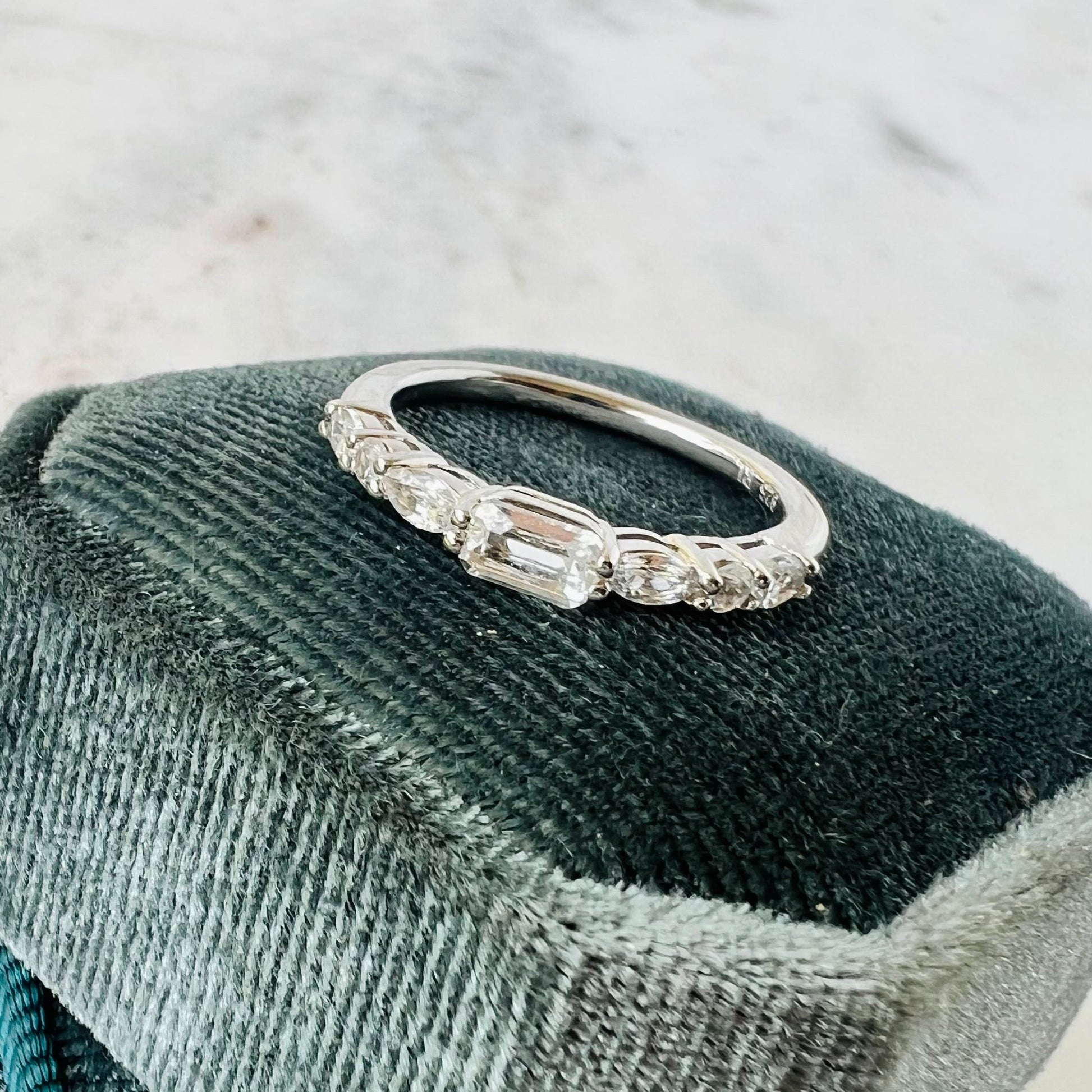 A silver band ring with emerald, oval and round-cut sapphire stones, displayed on a dark velvet jewelry cushion against a light marble background.