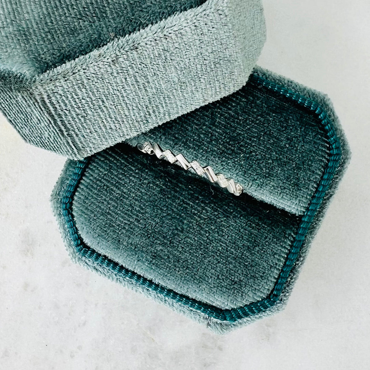 A silver ring with a textured zigzag pattern, displayed inside an open teal velvet ring box on a light marble surface.
