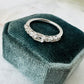A silver band ring with clear emerald, oval and round-cut stones, displayed on a dark velvet jewelry box over a light marble surface.