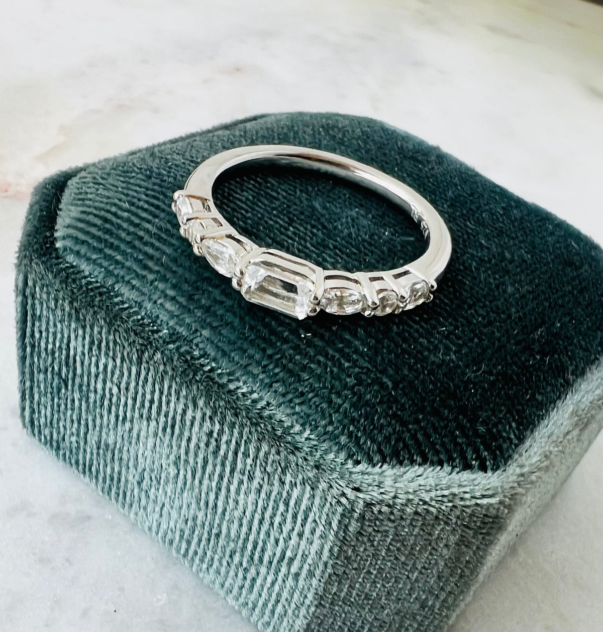 A silver band ring with clear emerald, oval and round-cut stones, displayed on a dark velvet jewelry box over a light marble surface.