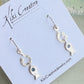 A pair of silver fish drop earrings with small dark green stones, displayed on a white Kiki Creative jewelry card.