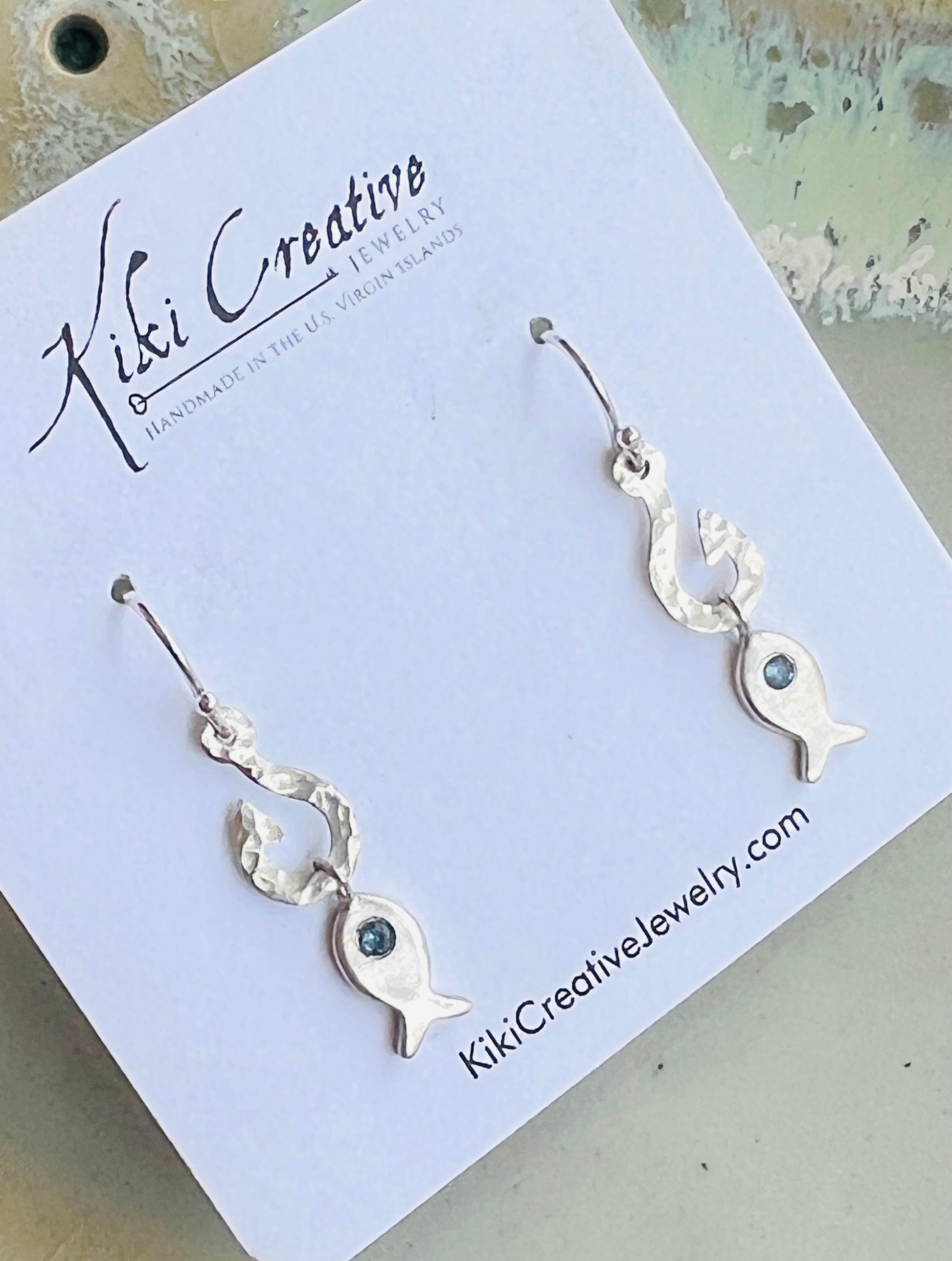 A pair of silver fish drop earrings with small dark green stones, displayed on a white Kiki Creative jewelry card.