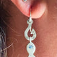 A close-up of a person's ear wearing a silver fish-shaped dangle earring with a small blue gemstone, alongside another stud earring.