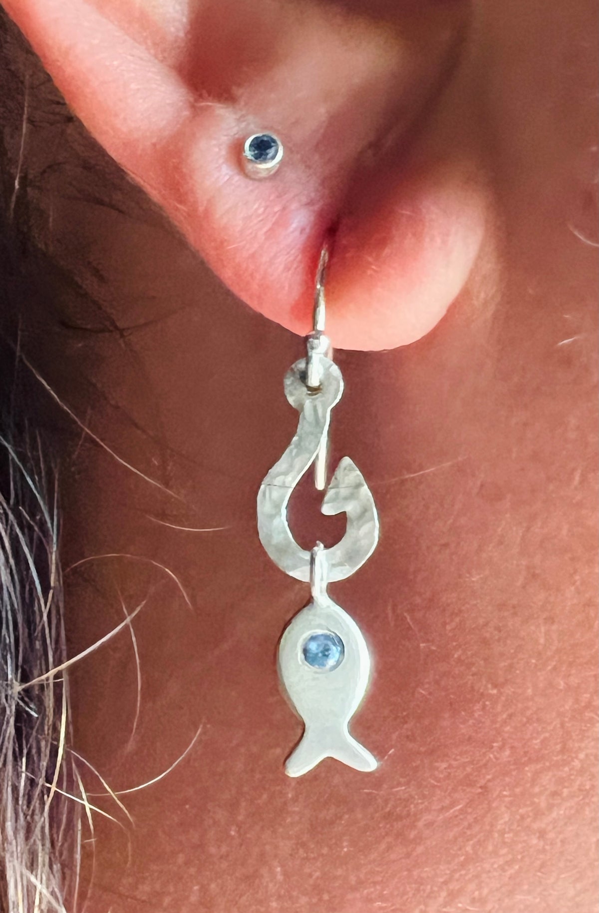 A close-up of a person's ear wearing a silver fish-shaped dangle earring with a small blue gemstone, alongside another stud earring.