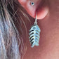 A close-up of a person's ear wearing a silver fish skeleton dangle earring, with a small blue gemstone stud earring above it.