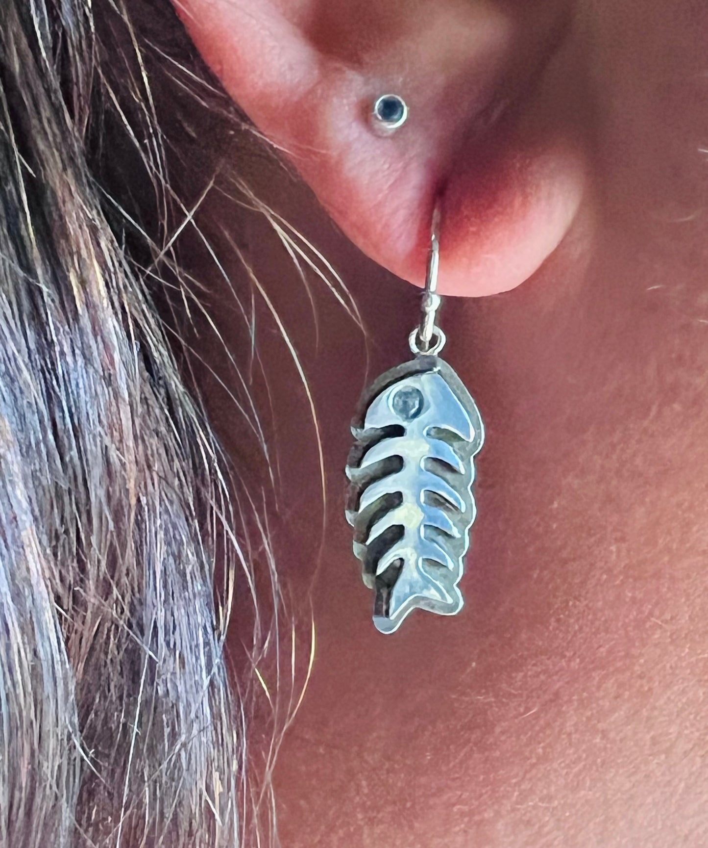 A close-up of a person's ear wearing a silver fish skeleton dangle earring, with a small blue gemstone stud earring above it.