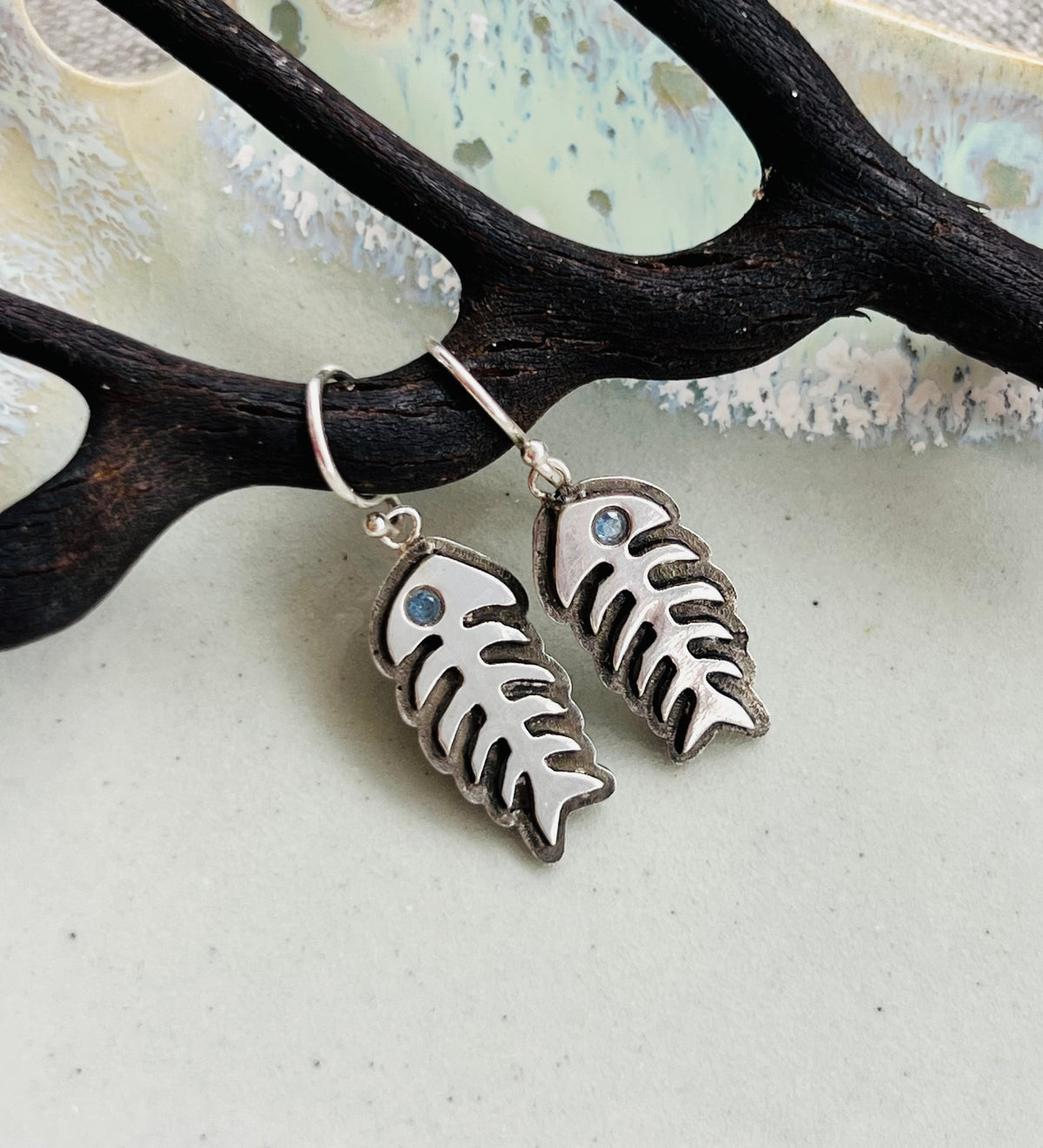 A pair of silver fish skeleton drop earrings with small blue stone accents, displayed on a dark branch against a light textured background.