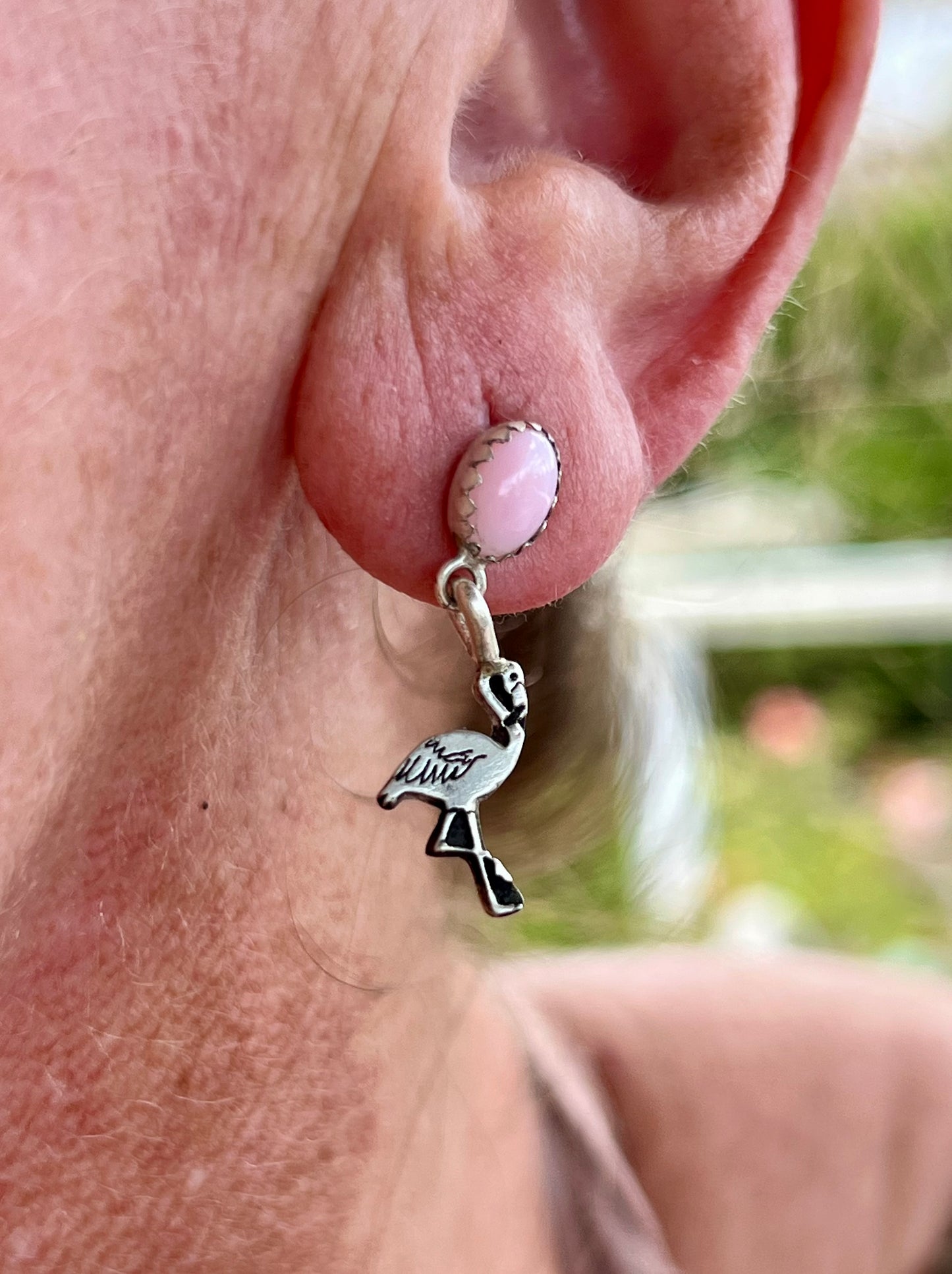 Close-up of an ear wearing a pink stone and flamingo charm earring with a blurred outdoor background.