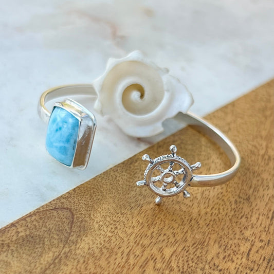 Silver ring with blue gemstone and ship's wheel design on a wooden surface with a shell.