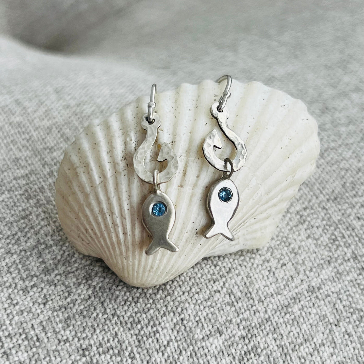 A pair of silver fish drop earrings with small blue gemstone accents, displayed on a white seashell against a textured gray fabric background.