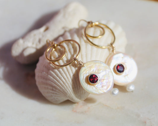 Gold hoop earrings with round iridescent shell pendants and small dark red gemstone centers, displayed on white seashells against a light stone surface.