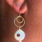 A close-up of a person's ear wearing a gold hoop earring with a dangling white iridescent bead and a small red gemstone accent.
