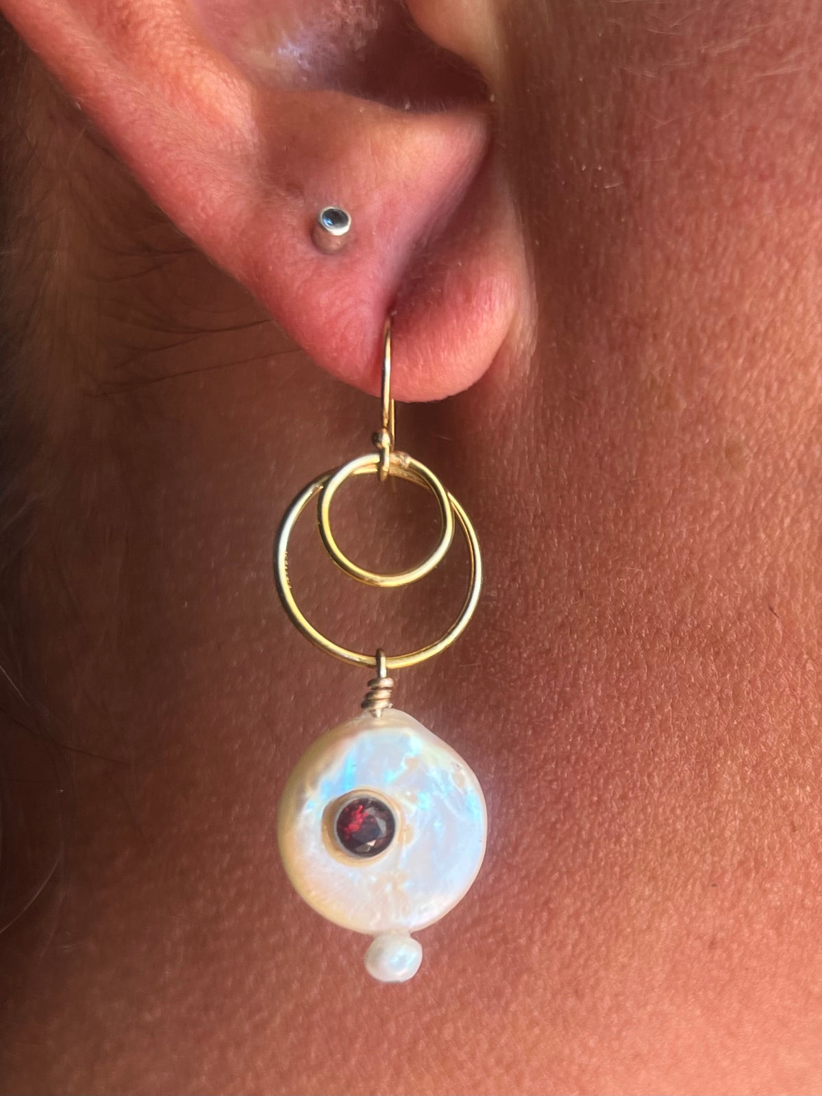 A close-up of a person's ear wearing a gold hoop earring with a dangling white iridescent bead and a small red gemstone accent.