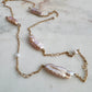 A gold chain necklace featuring irregular baroque pearls in soft pink and cream tones, accented with small white beads, laid out on a light marble surface.