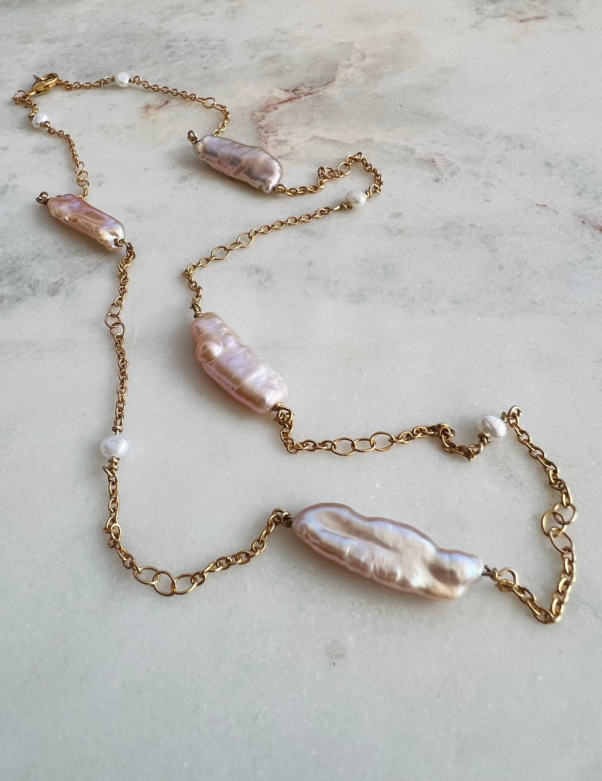 A gold chain necklace featuring irregular baroque pearls in soft pink and cream tones, accented with small white beads, laid out on a light marble surface.