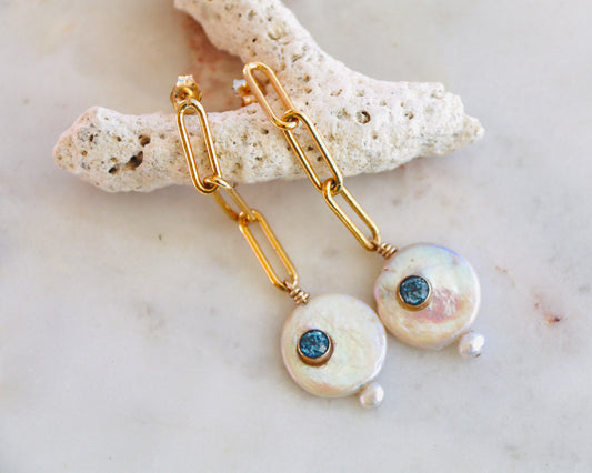 A pair of gold chain drop earrings with round iridescent shell pendants, each featuring a small dark blue stone and a tiny pearl bead, displayed on a light stone branch over a marble surface.