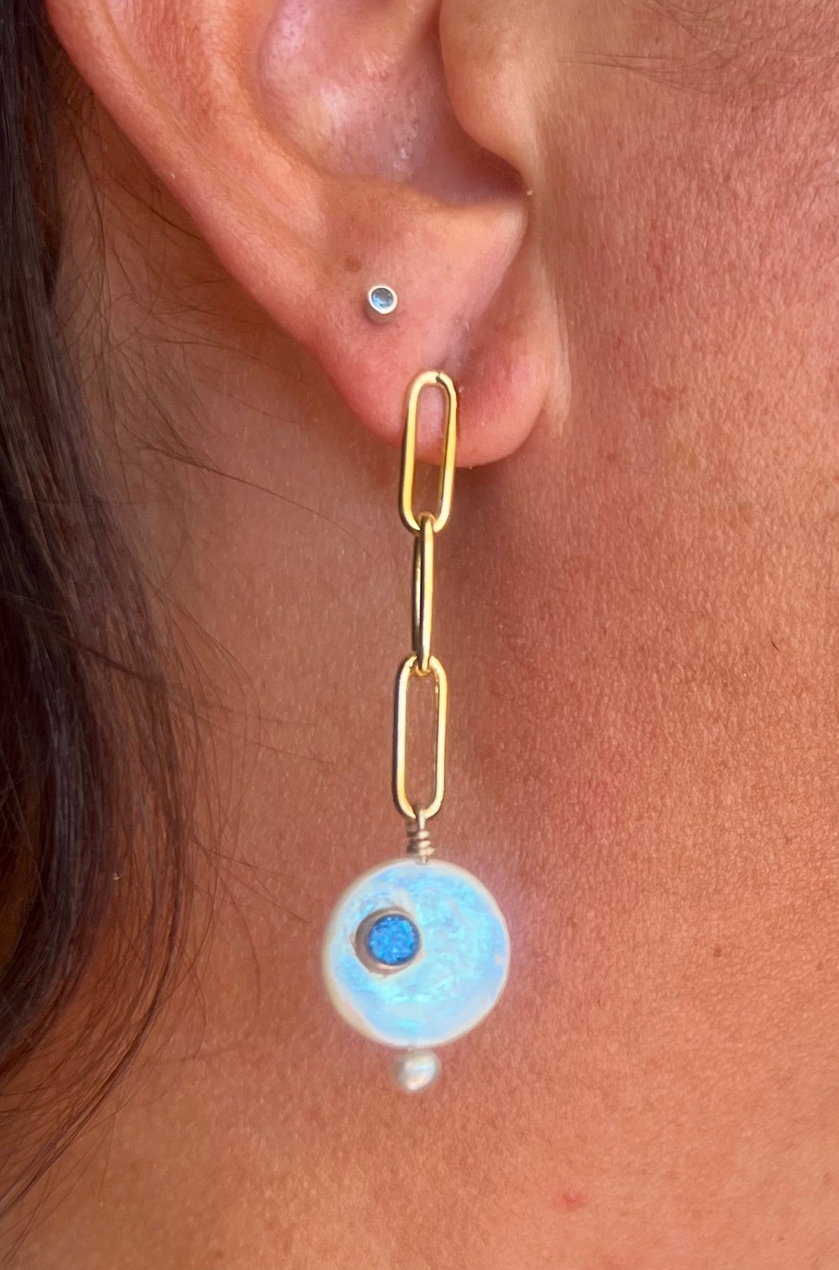 A close-up of a person's ear wearing a gold paperclip drop earring with a light blue circular disc and a small blue center, alongside a small silver stud earring.