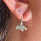 A close-up of a person's ear wearing a silver huggie hoop earring with a dangling map-shaped charm featuring a swirl symbol, alongside a small silver stud piercing.