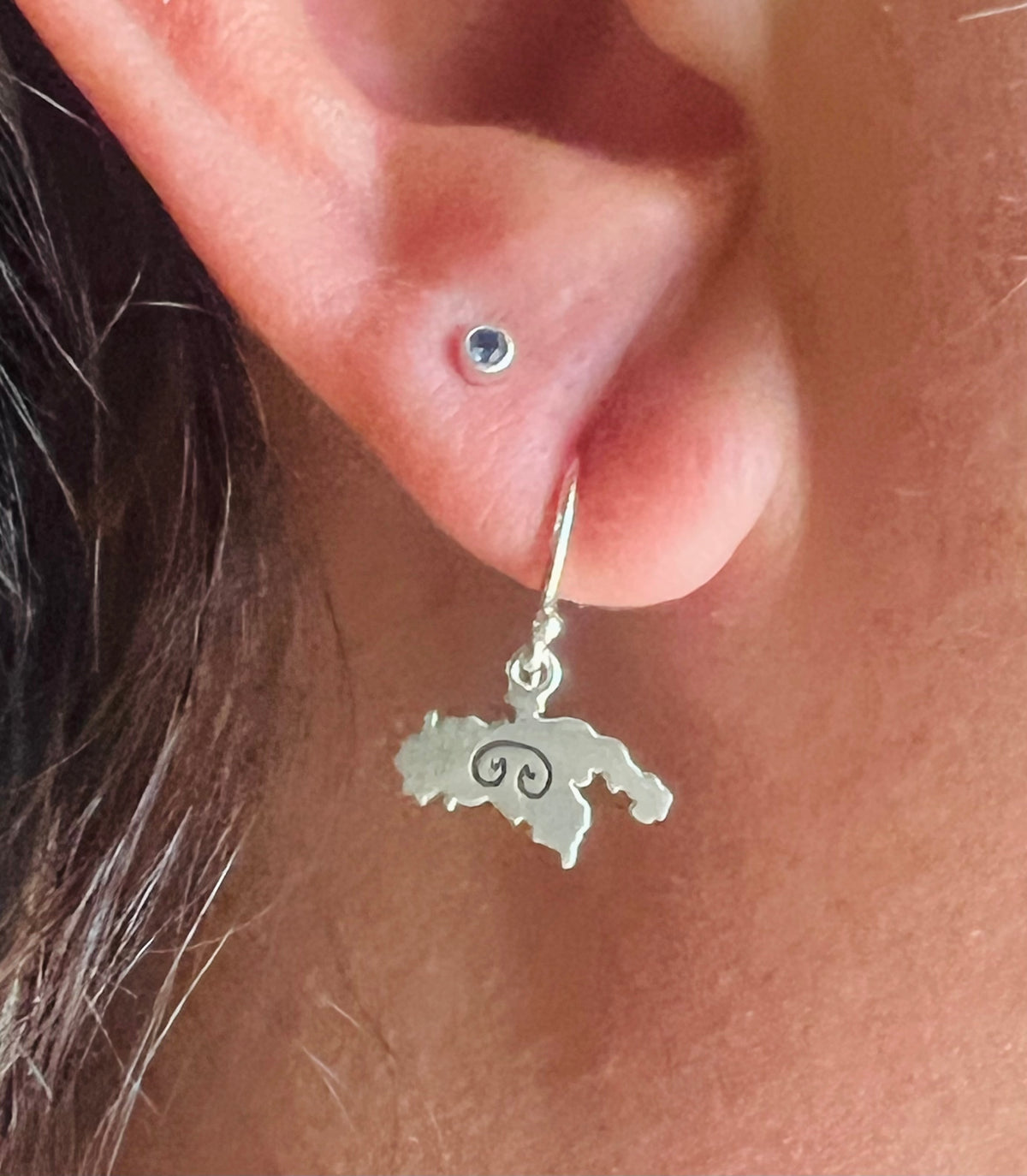 A close-up of a person's ear wearing a silver huggie hoop earring with a dangling map-shaped charm featuring a swirl symbol, alongside a small silver stud piercing.