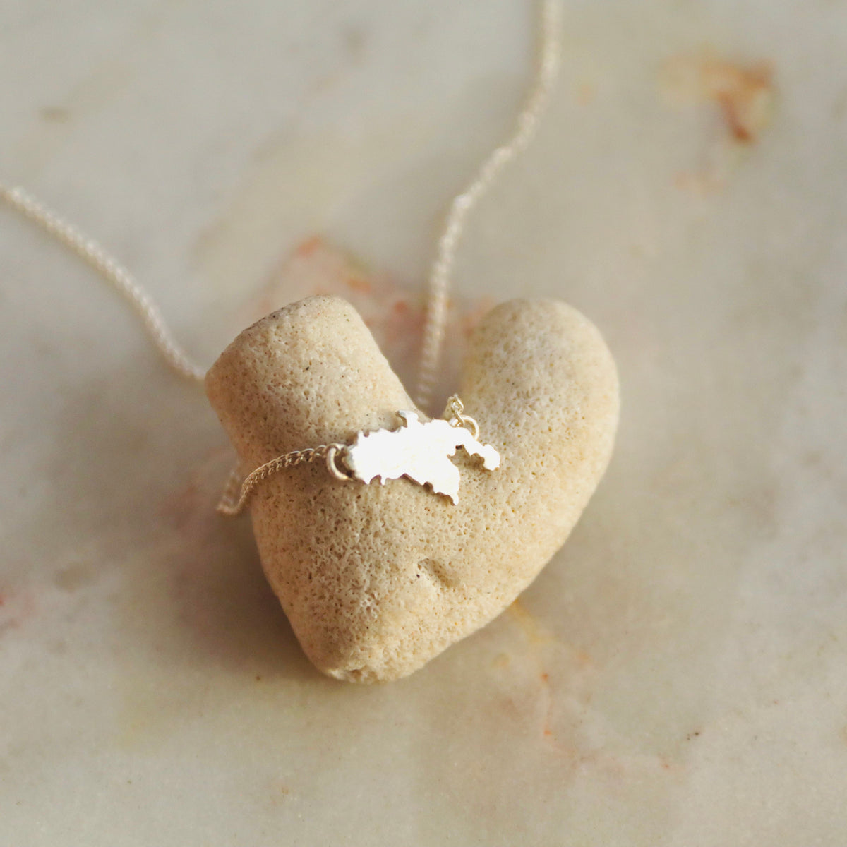 A heart-shaped pendant necklace made from a small beige stone or coral piece, with a delicate silver map-shaped charm attached, resting on a light beige marble surface.