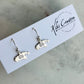 A pair of silver drop earrings shaped like the state of Michigan with a swirl design inside, displayed on a white brand card for Kiki Creative Jewelry on a light marble surface.