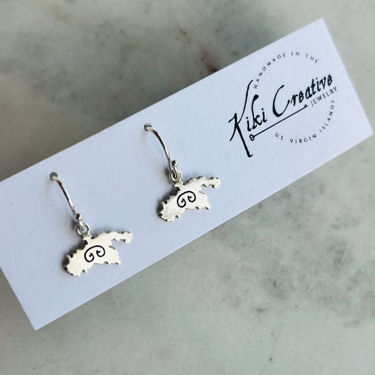 A pair of silver drop earrings shaped like the state of Michigan with a swirl design inside, displayed on a white brand card for Kiki Creative Jewelry on a light marble surface.