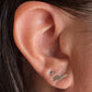 Close-up of a person's ear showing two small earrings: a tiny silver stud with a dark gemstone and a small silver flat piece on the lobe, with dark hair visible around the ear.