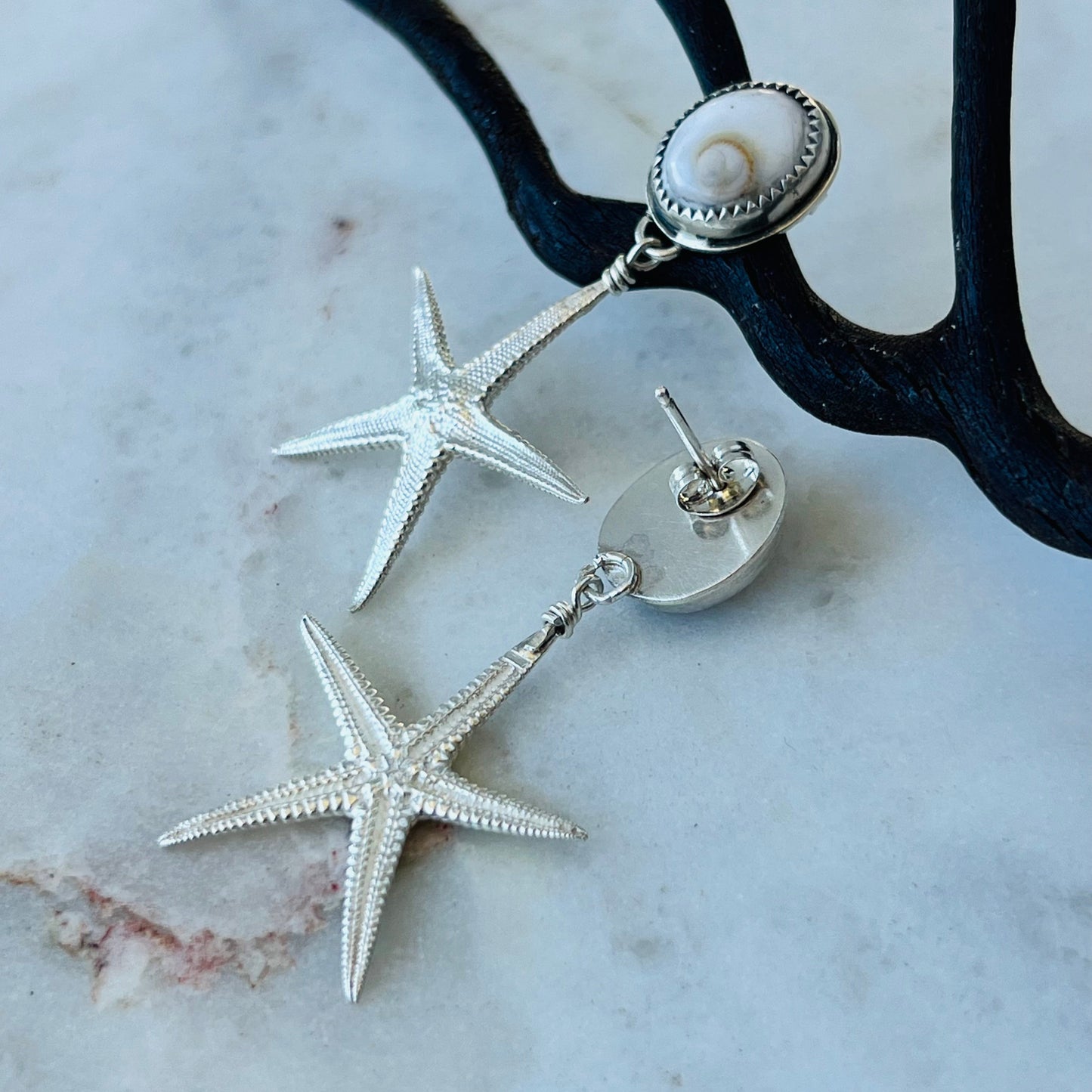 Silver starfish earrings on a marble surface with a black metal stand.