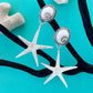 Starfish-shaped earrings with pearl-like centers on a turquoise background with black seaweed and white shells.