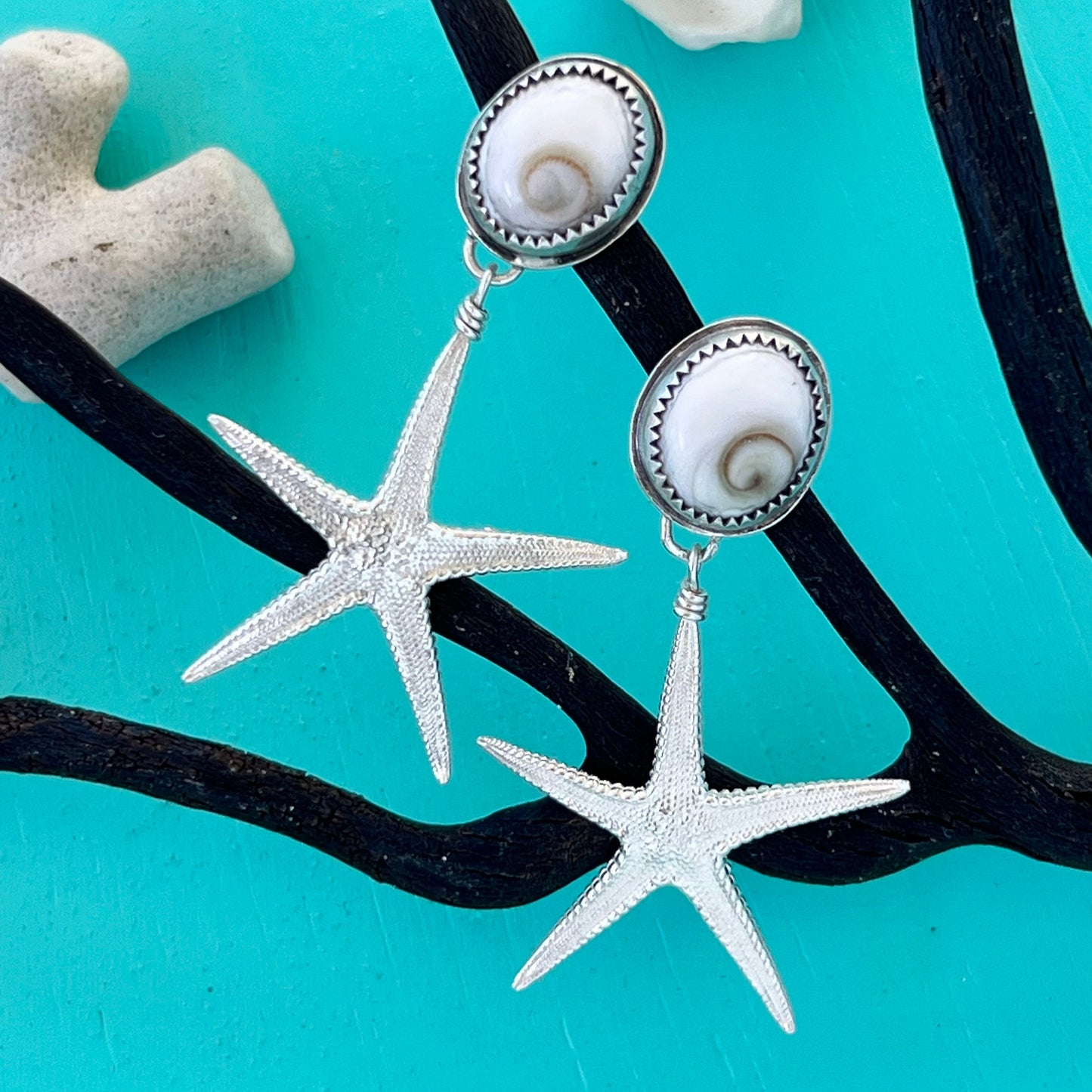Starfish-shaped earrings with pearl-like centers on a turquoise background with black seaweed and white shells.