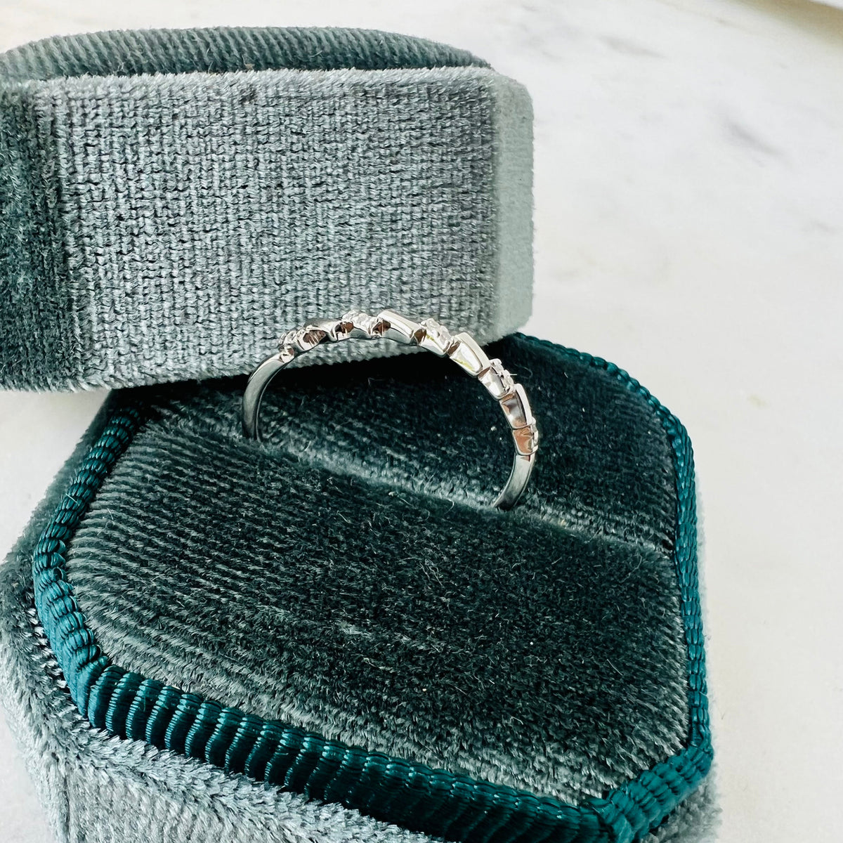 A silver band ring with small diamond stones, displayed on a dark teal velvet ring box with a matching lid on a light marble surface.