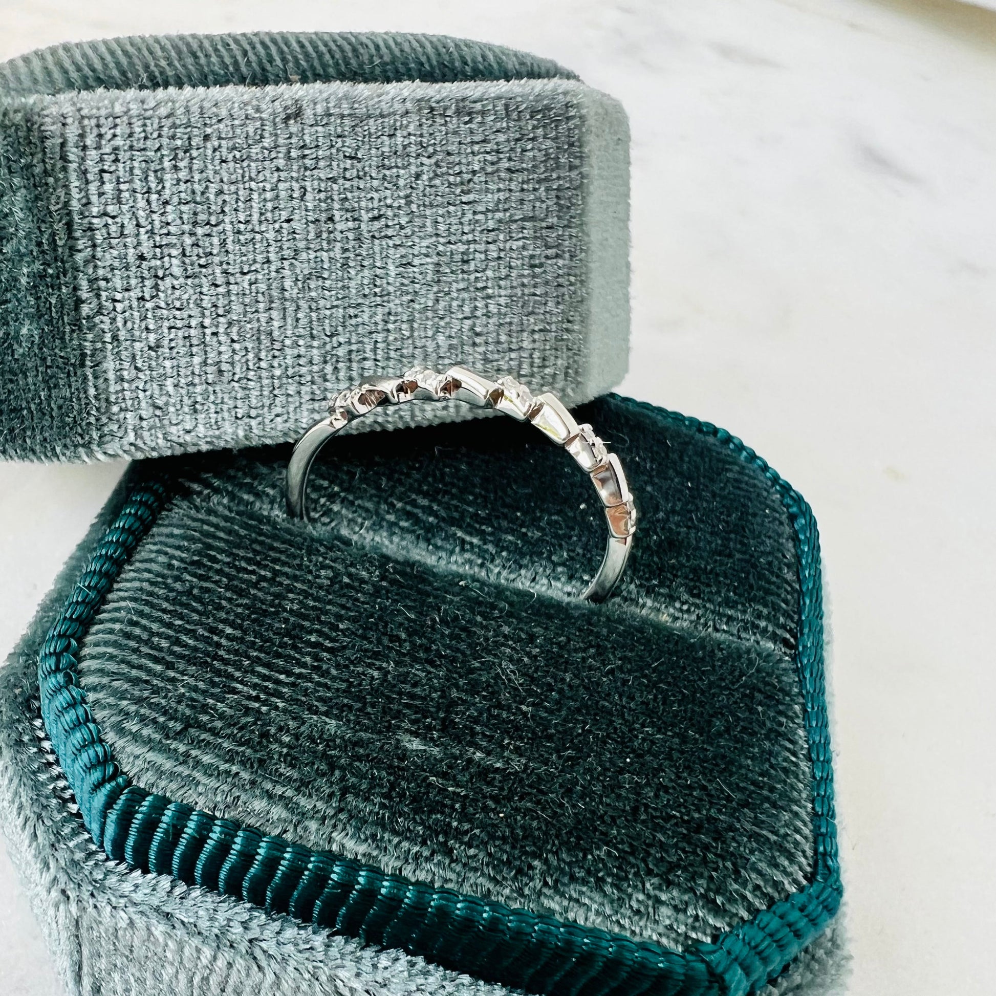 A silver band ring with small diamond stones, displayed on a dark teal velvet ring box with a matching lid on a light marble surface.