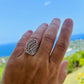 Hand wearing a silver ring with a patterned design against a scenic background