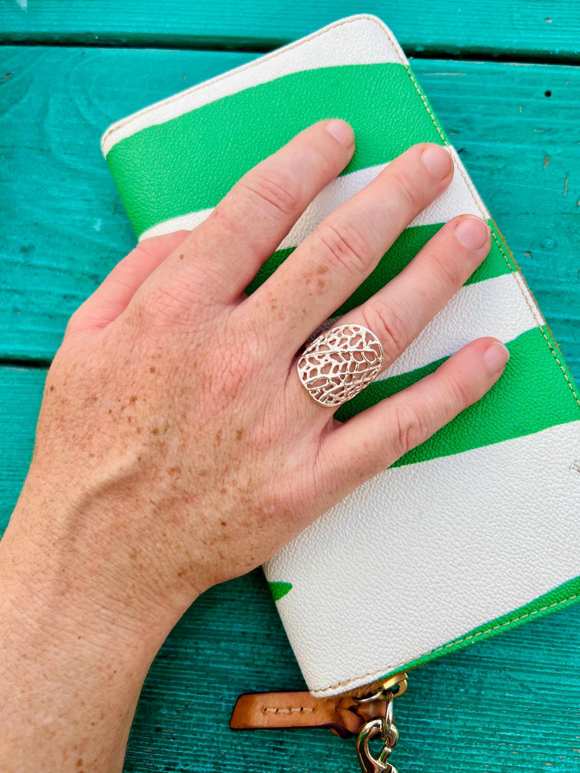 Hand holding a green and white clutch with a decorative ring on a teal wooden surface