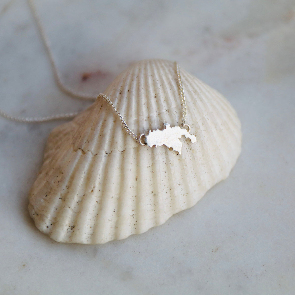 A silver necklace with a small map-shaped pendant resting on a large beige seashell, set against a light marble background.