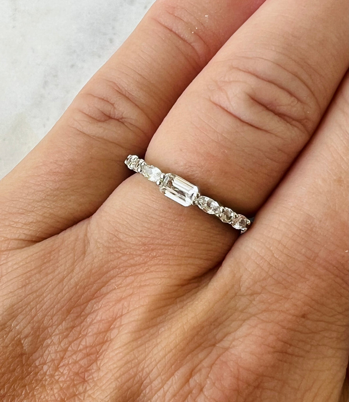 A close-up of a hand wearing a silver eternity ring with a central emerald-cut sapphire and smaller side sapphires.